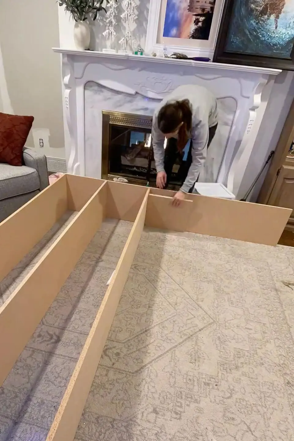 DIYer assembling a large wooden frame for built-in storage in the living room, using MDF panels near a fireplace.